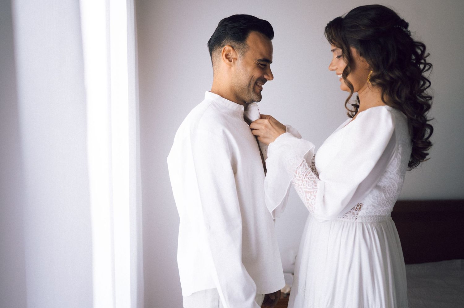 Couple in white outfits sharing a tender moment, woman adjusting man's collar near a bright window.