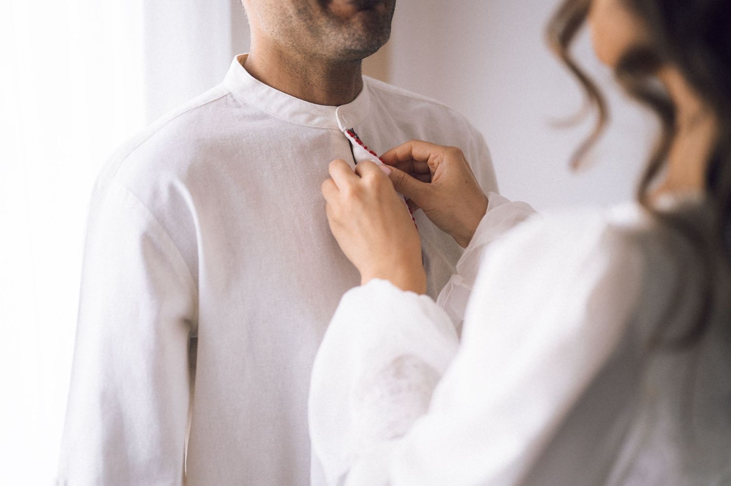 Woman in white adjusting a man's white kurta shirt collar during a wedding preparation ceremony.
