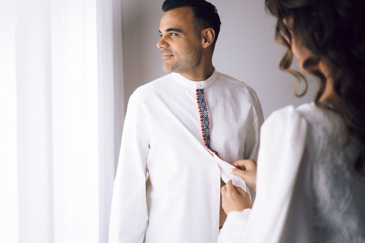 Woman helping man button white embroidered kurta shirt near bright window during wedding preparation.