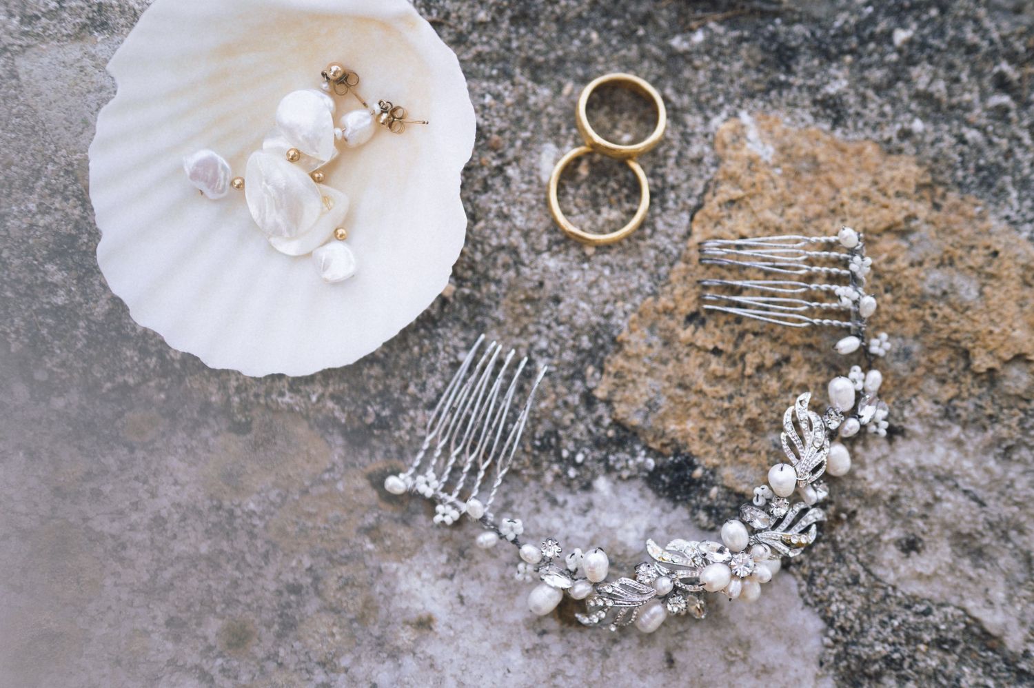 Bridal jewelry flatlay with pearl earrings, gold rings, crystal hair comb, and floral headpiece on stone surface.