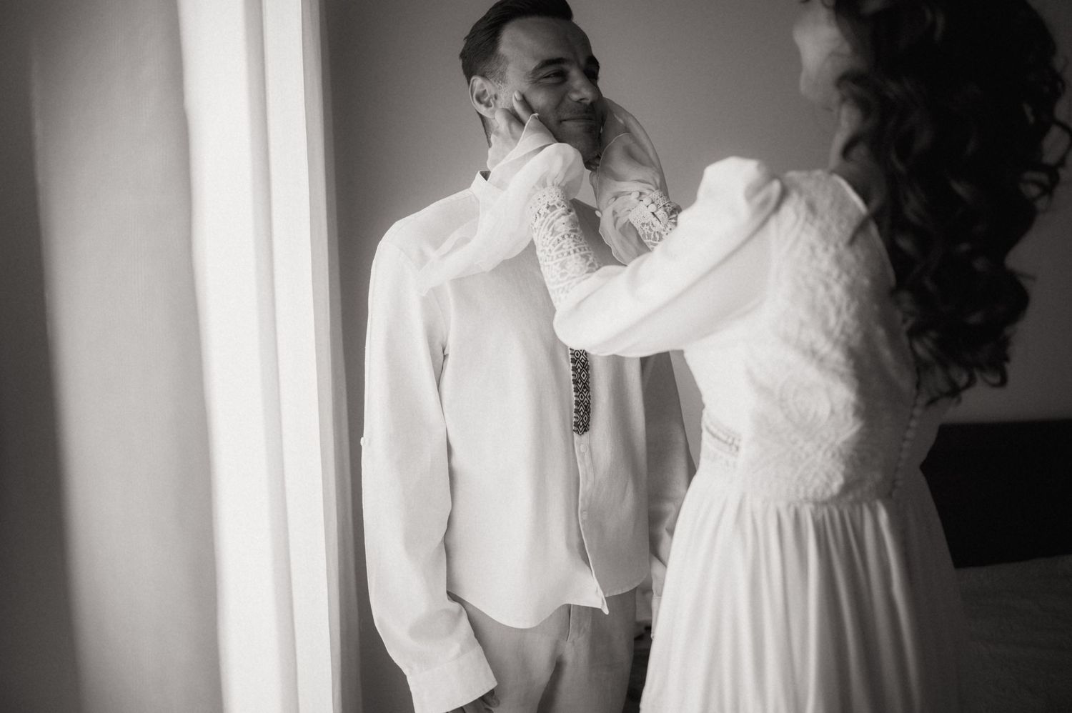 Black and white photo of a bride adjusting the groom's collar before their wedding ceremony.