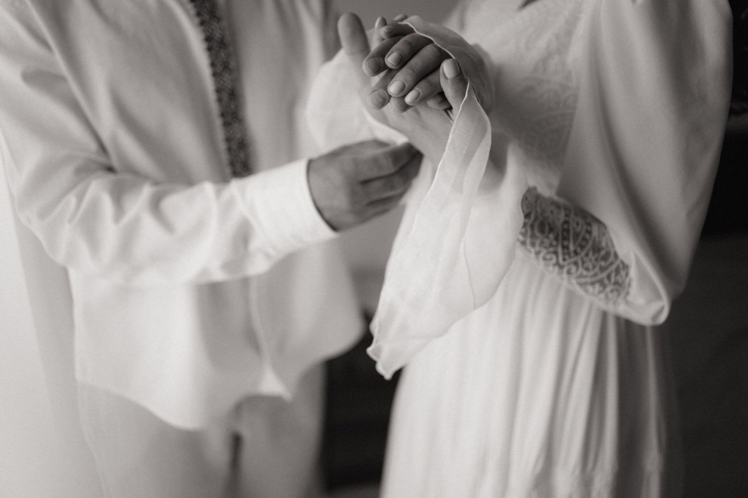 Black and white photo of two people in white garments holding hands during a wedding ceremony.