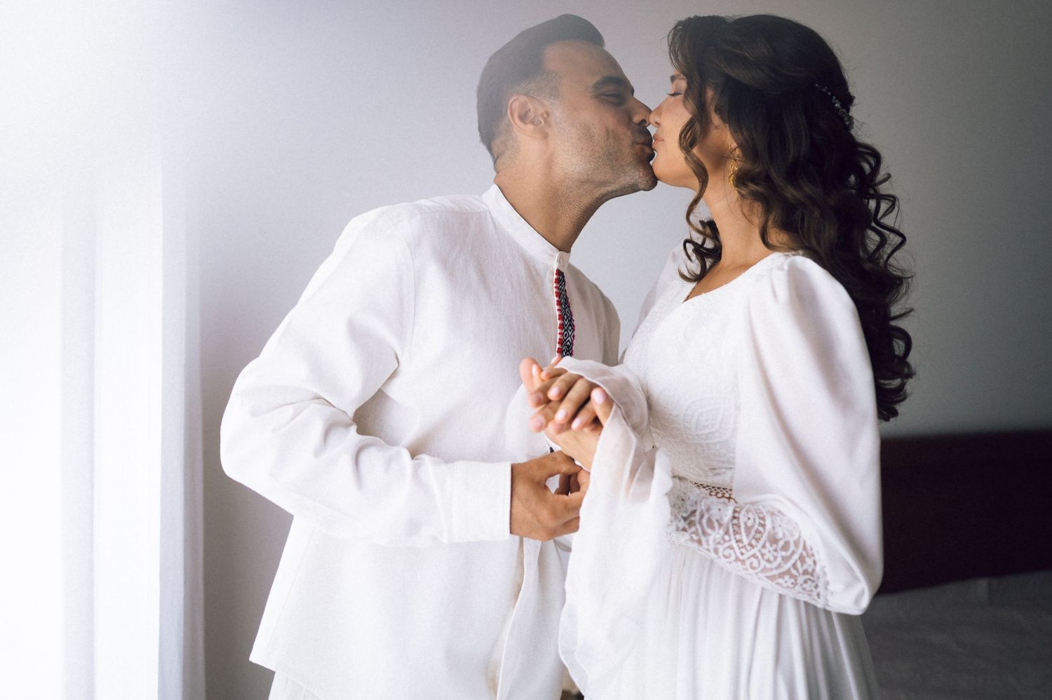 A couple in white attire shares a romantic kiss while holding hands in a bright, softly lit room.