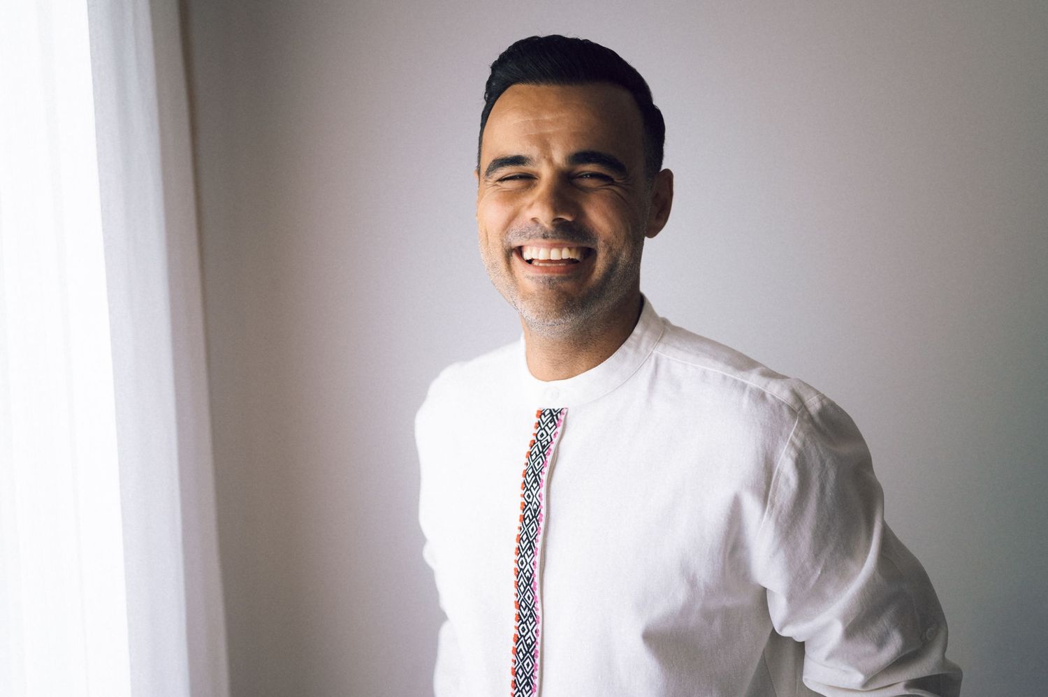 Smiling man in white shirt with colorful embroidered tie, posing confidently near a bright window.