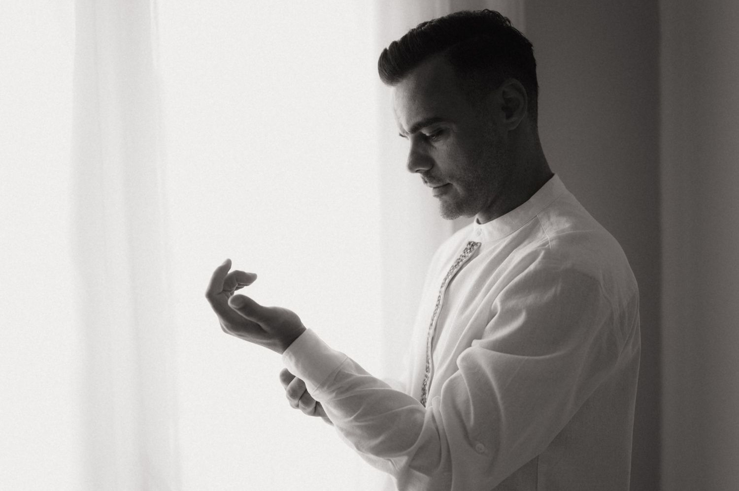 Black and white photo of a man in a white dress shirt adjusting his cufflinks by a bright window.