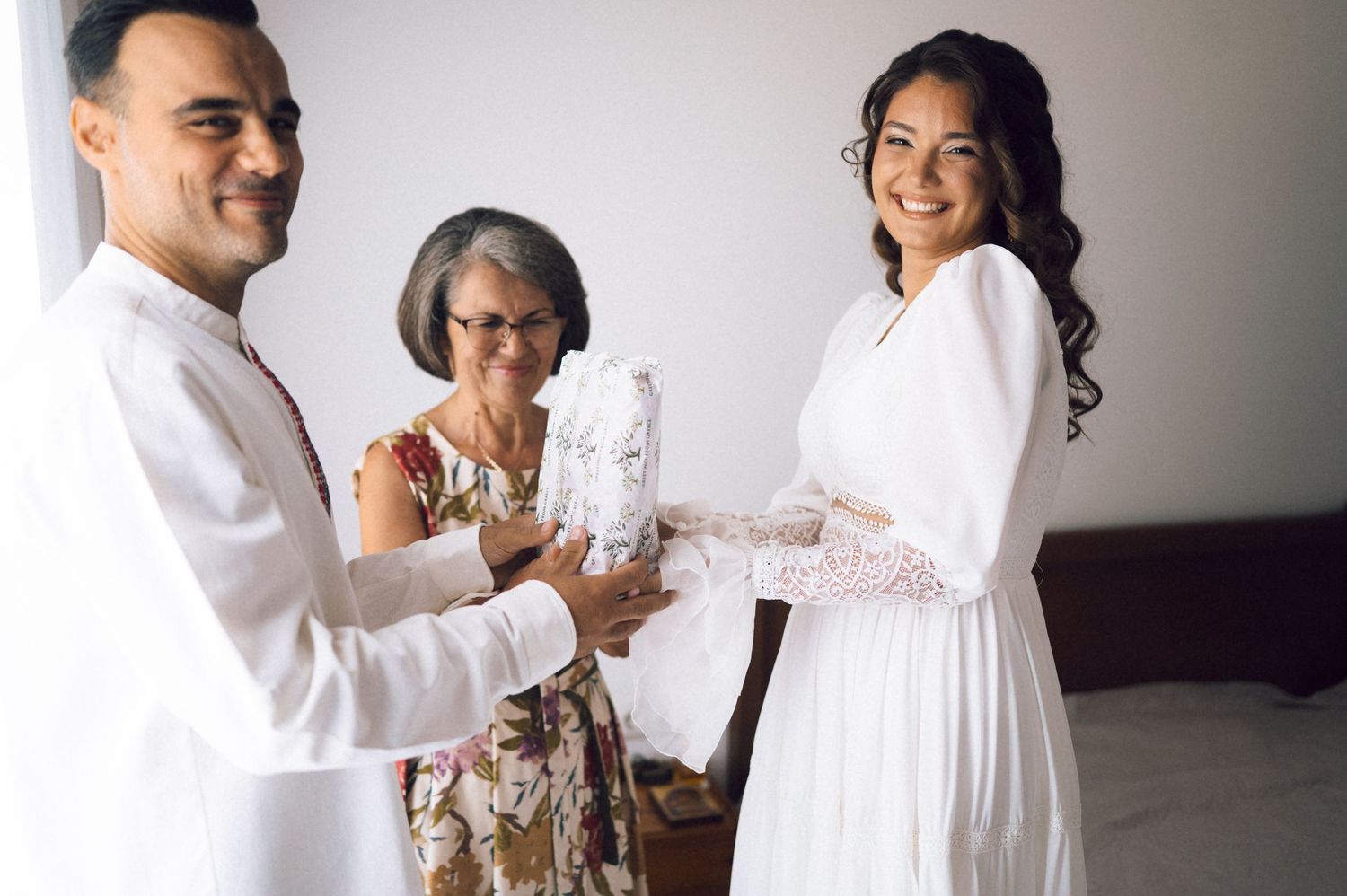 Smiling bride in white dress receiving a gift from groom with older woman watching during wedding celebration.