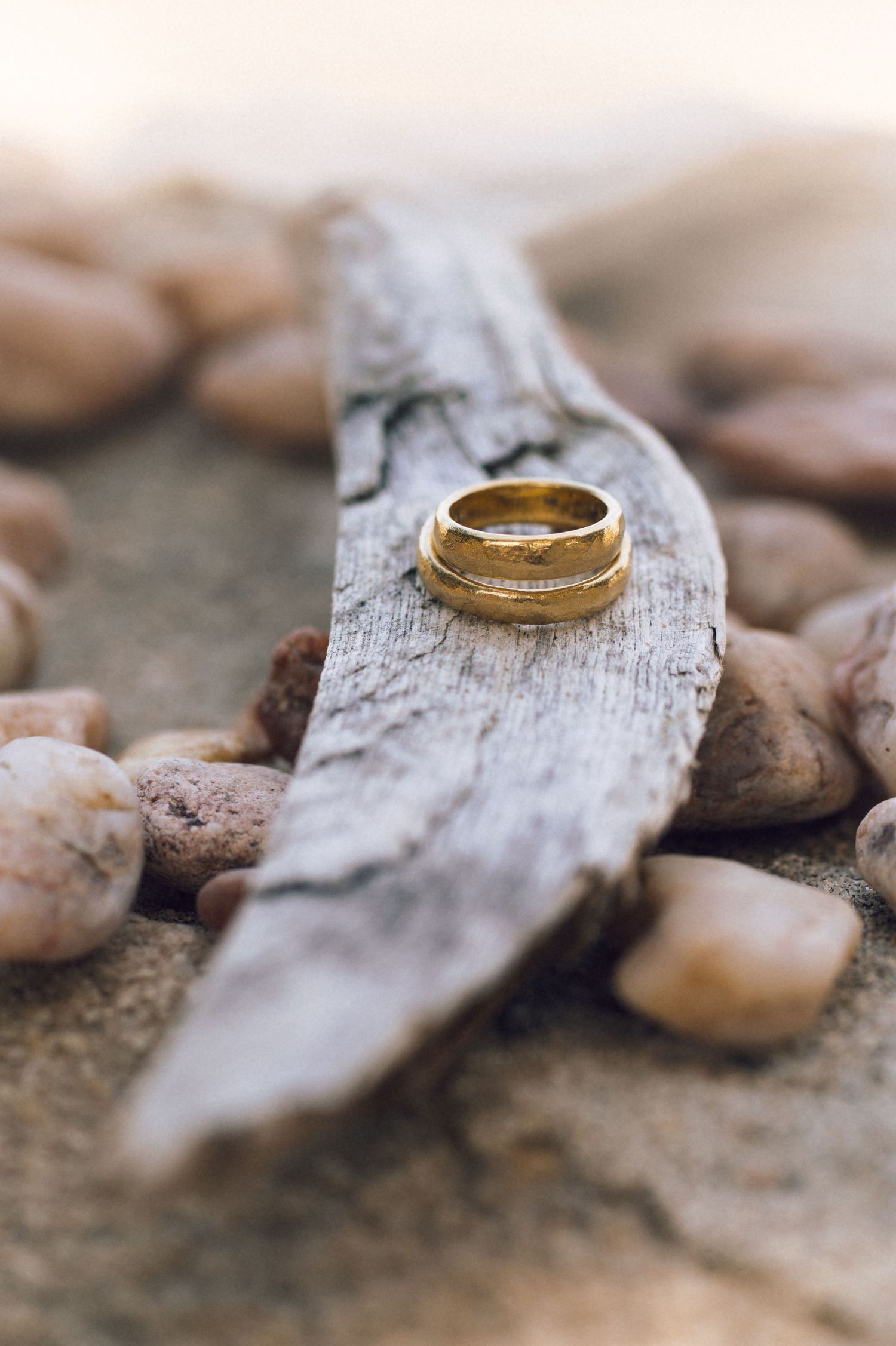 Two gold wedding rings stacked on driftwood surrounded by smooth pebbles, elegant rustic outdoor setting.