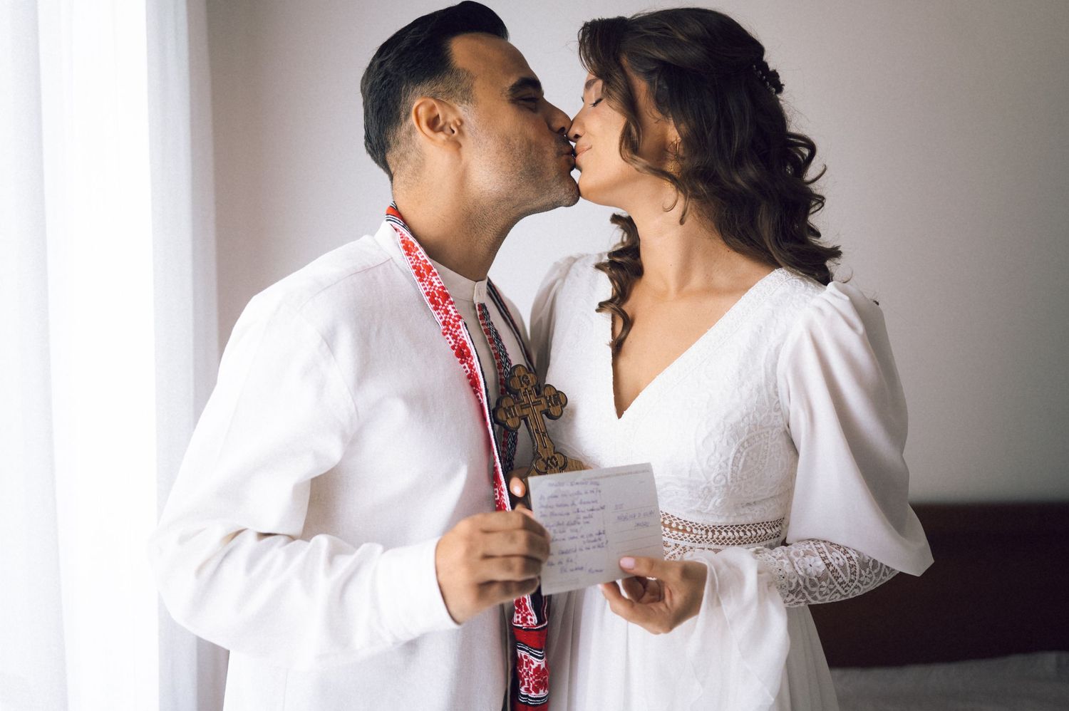 A couple in white wedding attire shares a kiss, the groom in an embroidered shirt and bride holding a card.
