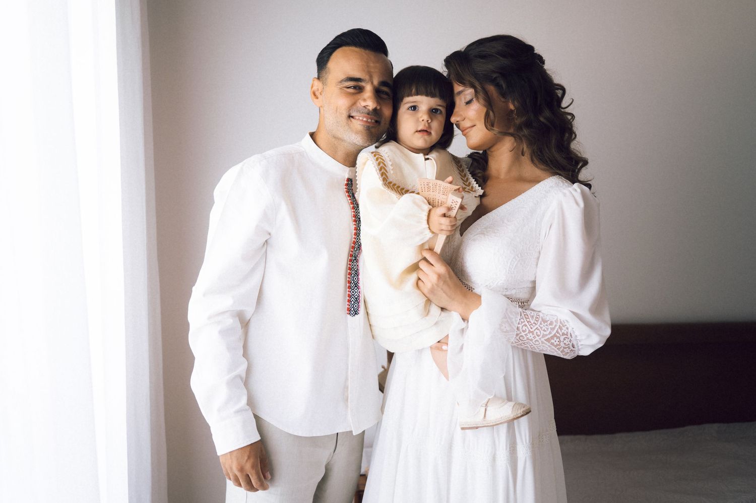 Happy family in white outfits, parents holding baby girl, posing together in bright room.