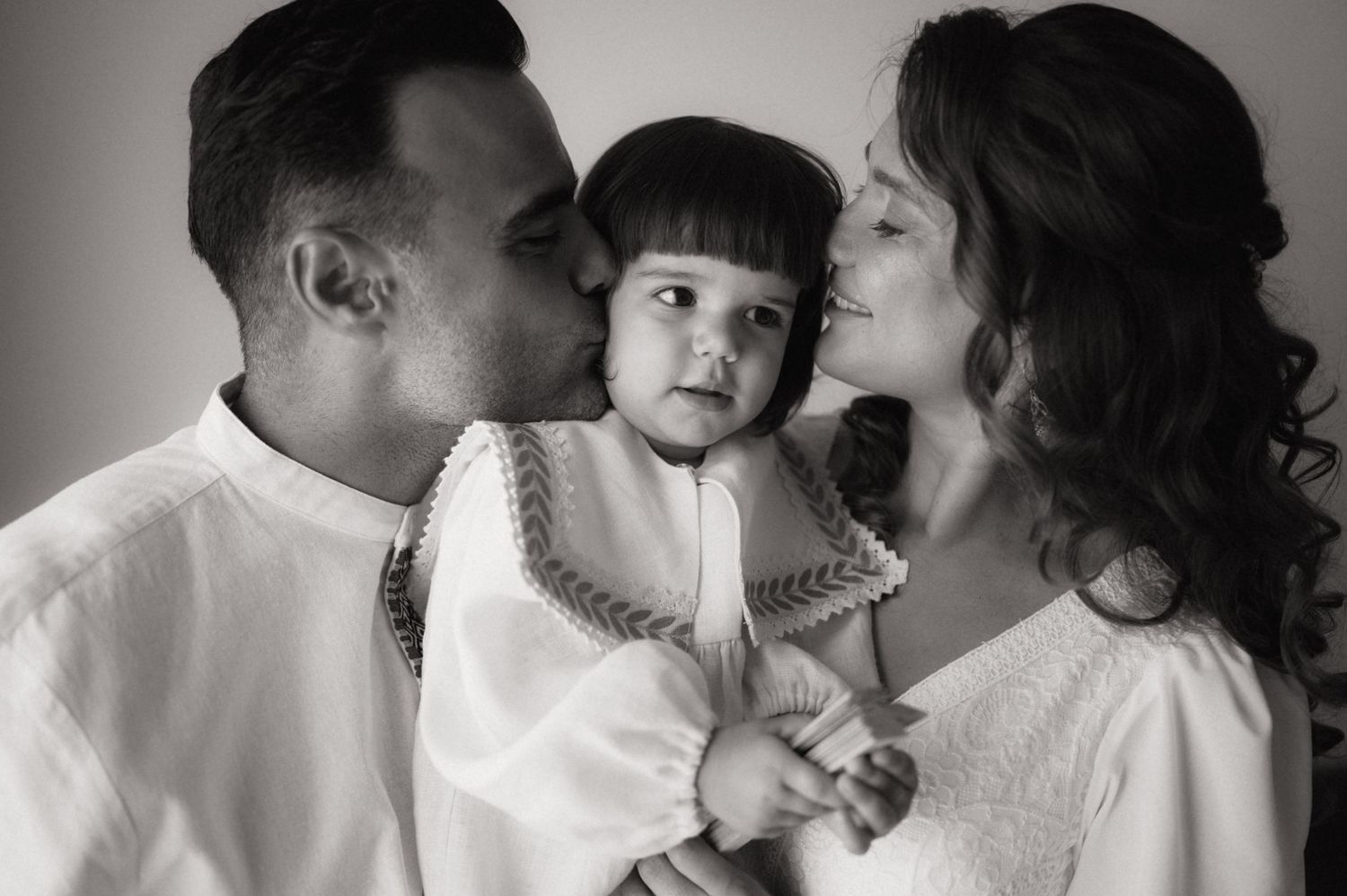 Black and white photo of parents kissing their toddler on cheeks, family portrait with warm expressions.
