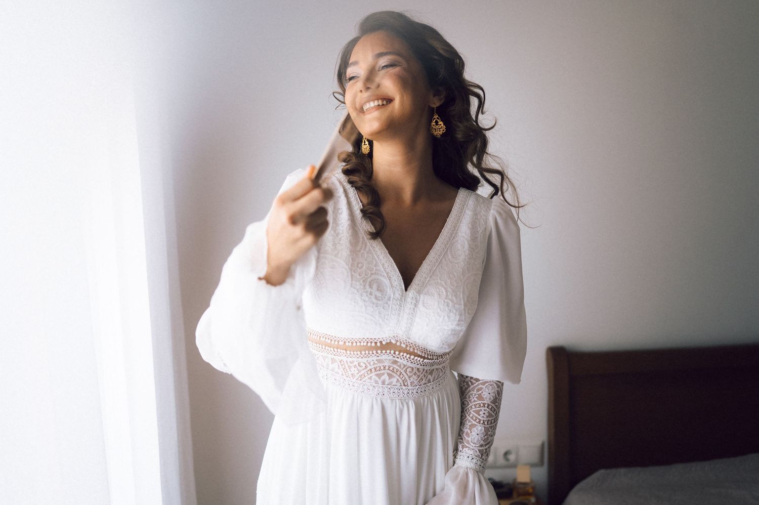 Smiling bride in a white boho wedding dress with lace details, posing by a bright window on her wedding day.