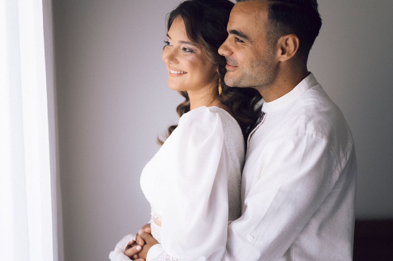 A happy couple in white outfits sharing a tender embrace near a bright window.