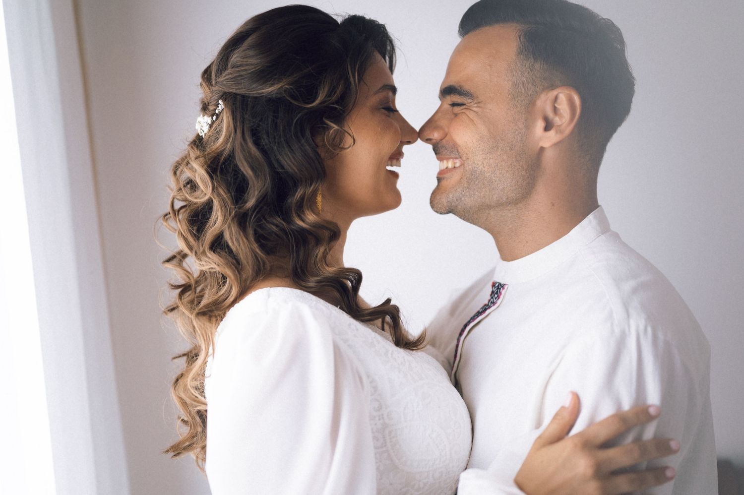 A smiling couple in white outfits share a romantic moment, gazing at each other lovingly indoors.