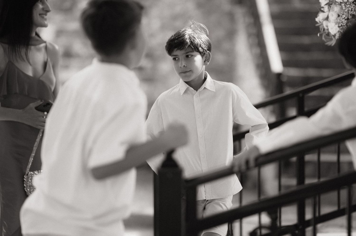 Black and white photo of a young boy in a white shirt standing near railing at an outdoor event.