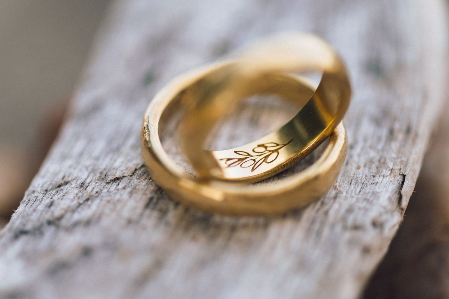Two gold wedding rings resting on rustic wood, one engraved with a delicate leaf design.