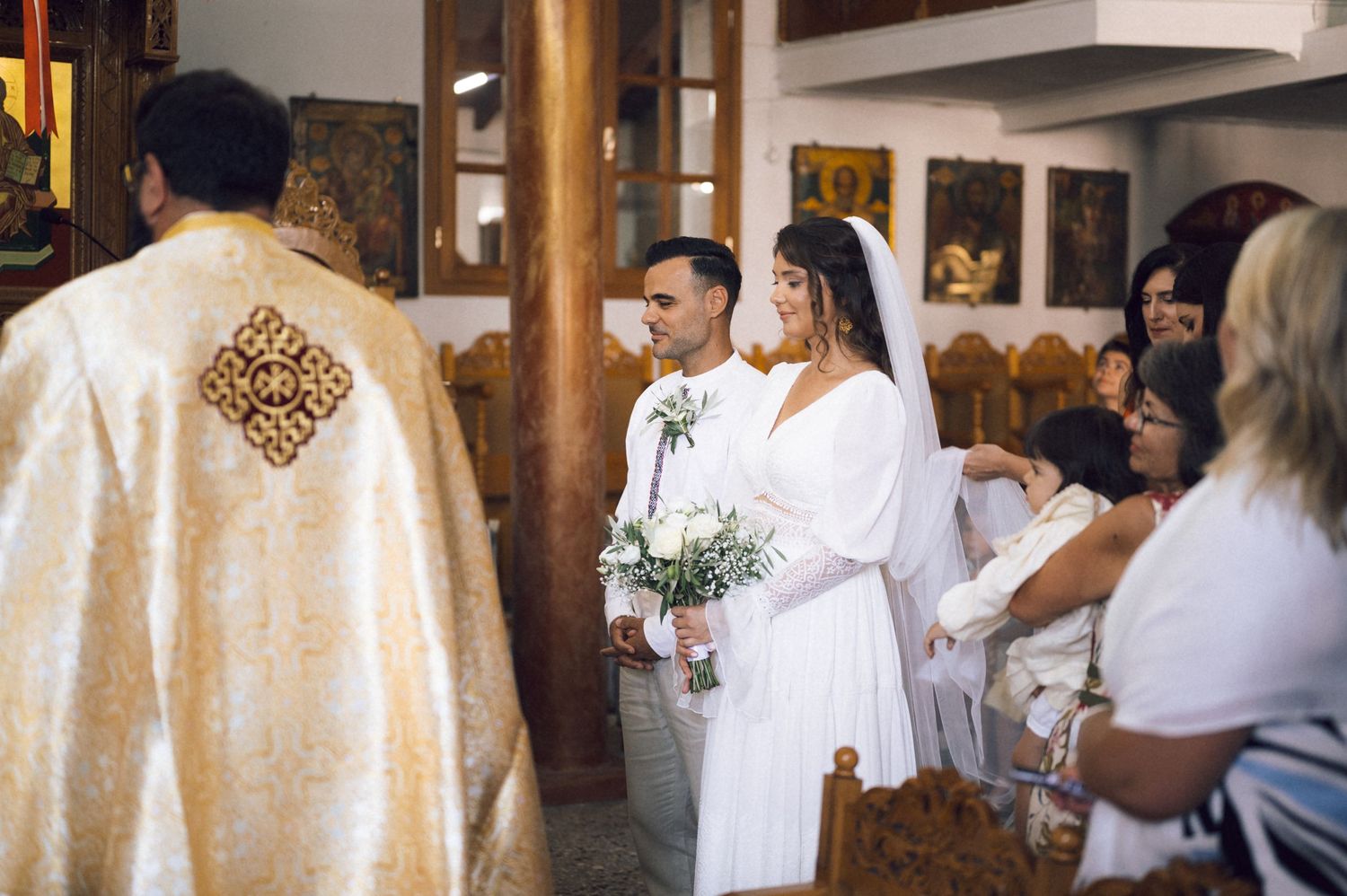 Bride and groom stand at the altar during an Orthodox church wedding ceremony, bride holding white floral bouquet.