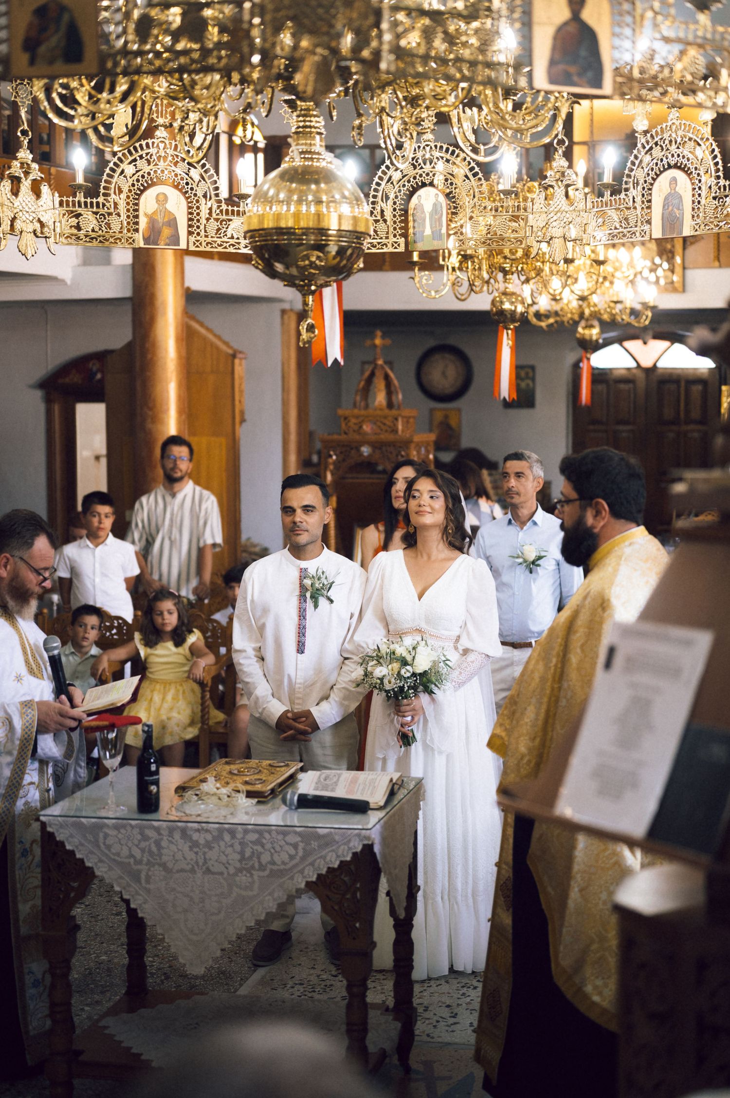 Couple exchanges vows in ornate Orthodox church, bride in white gown holding bouquet, groom in white suit, priest officiating.