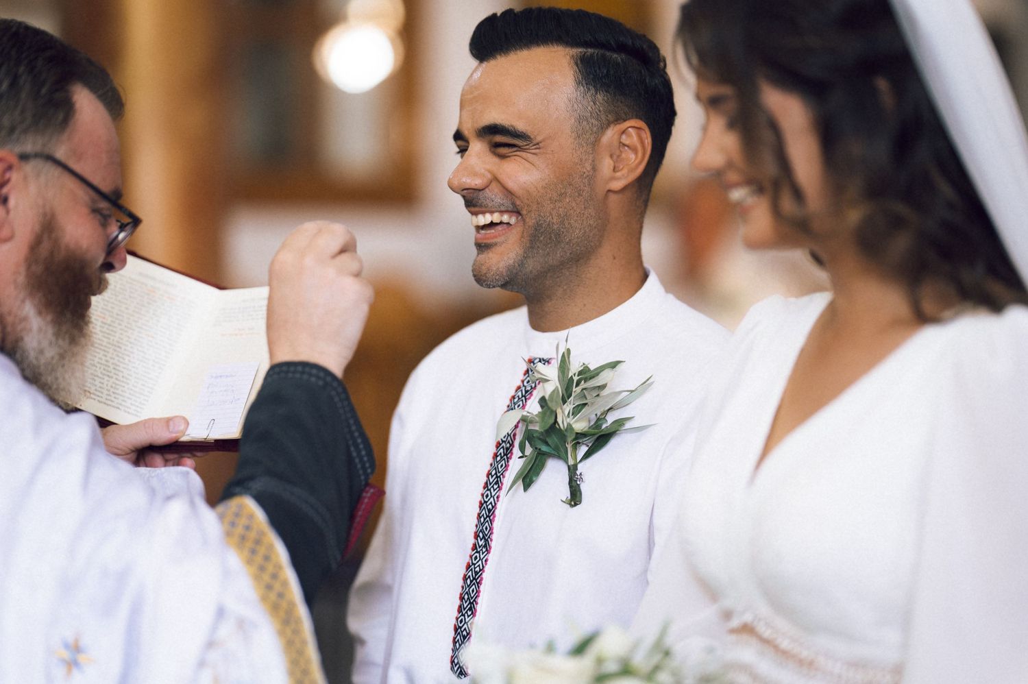 Groom smiling joyfully during wedding ceremony as priest reads scripture, bride in white dress beside him.