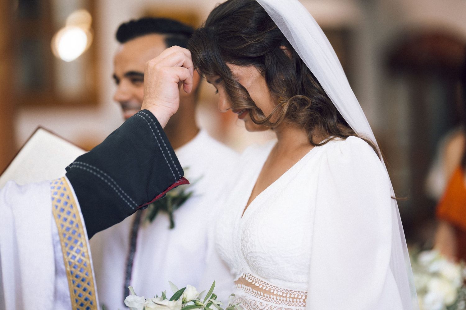Bride in white dress and veil receives blessing from priest during church wedding ceremony.