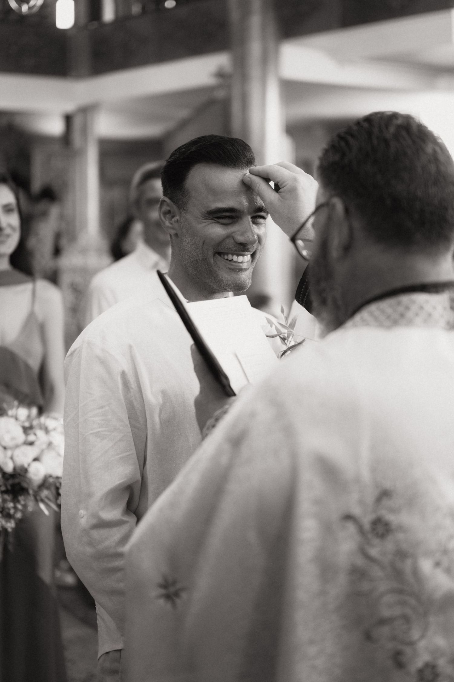 Black and white photo of a smiling groom receiving a blessing from a priest during a wedding ceremony.