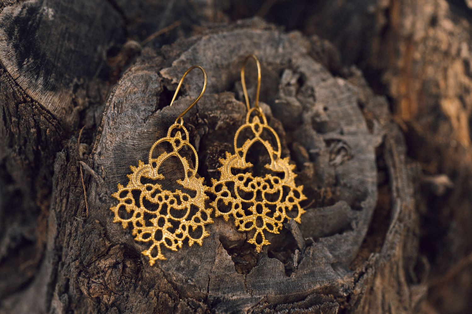 Ornate gold filigree drop earrings with intricate lace-like floral design displayed on rustic wood background.