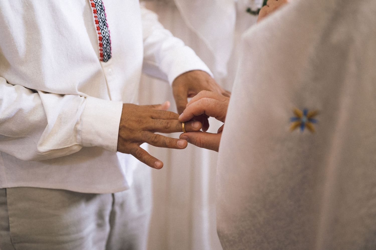 Close-up of two people exchanging wedding rings during a ceremony, wearing white embroidered traditional attire.