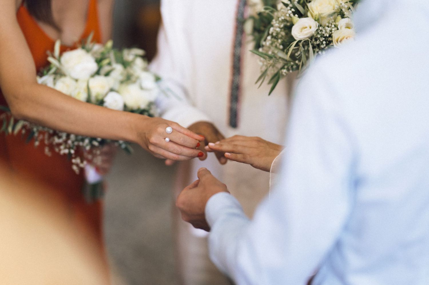 Couple exchanging wedding rings during ceremony, bride holding white floral bouquet with greenery.