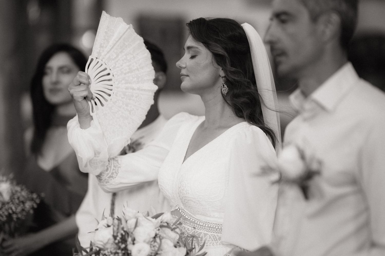 Elegant bride in white gown holding a decorative fan and bouquet at a wedding ceremony, black and white photo.