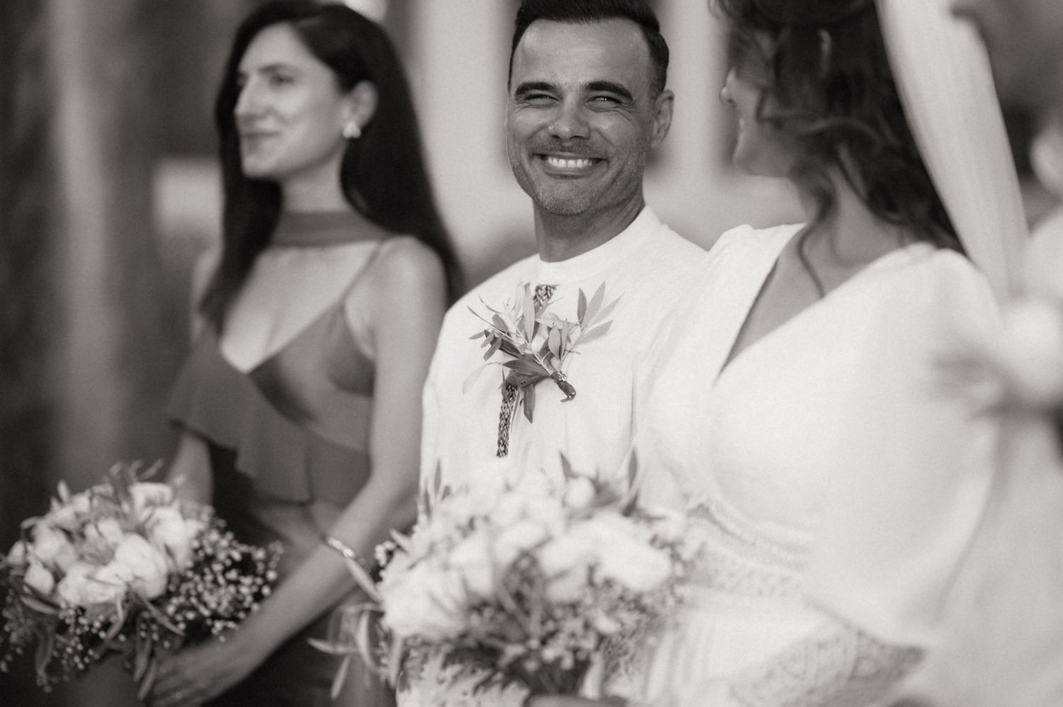 Black and white wedding photo of smiling groom with bridesmaids holding bouquets at elegant ceremony.