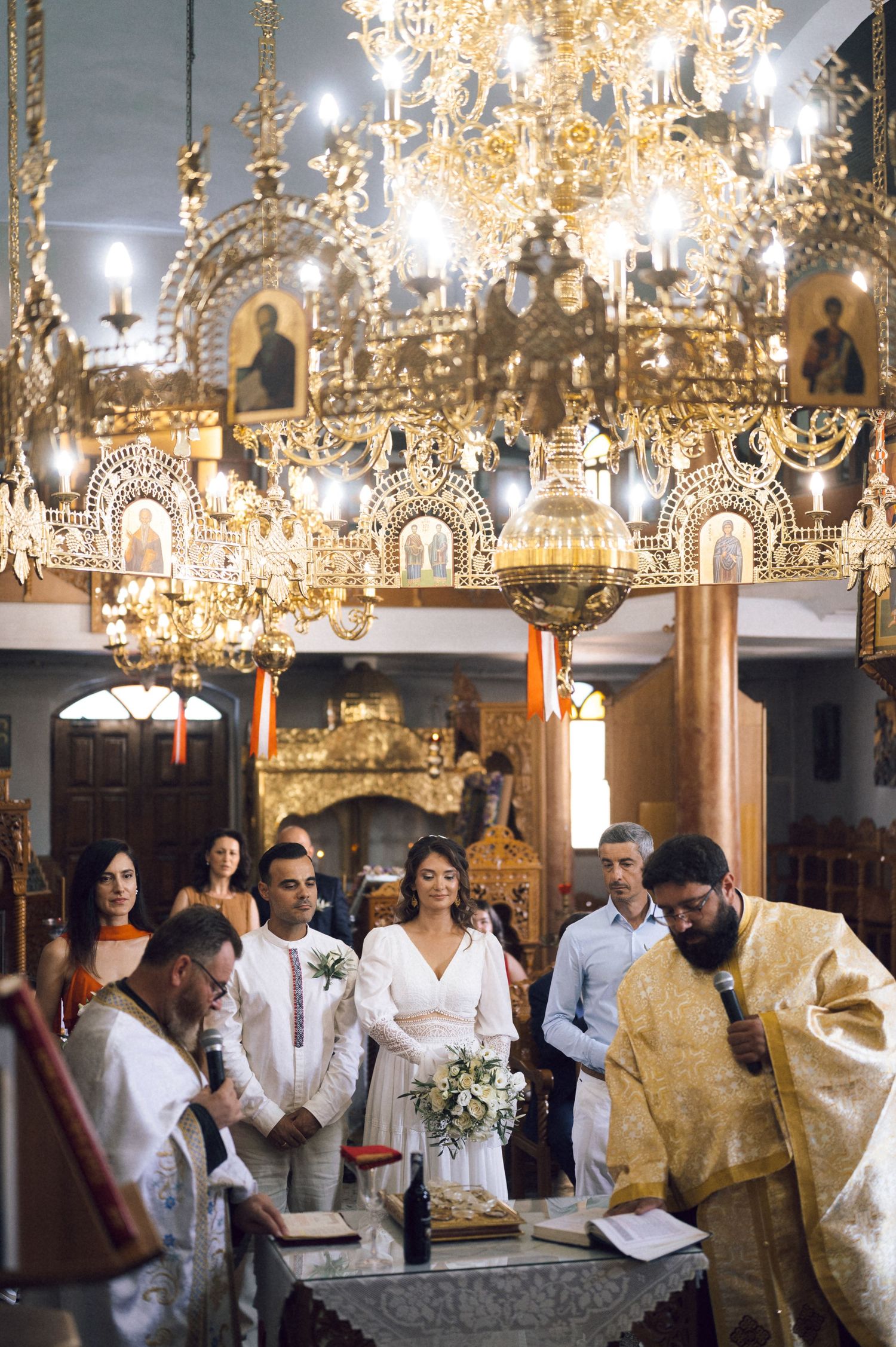 Greek Orthodox wedding ceremony with bride and groom standing before a priest under ornate golden chandeliers in a church.