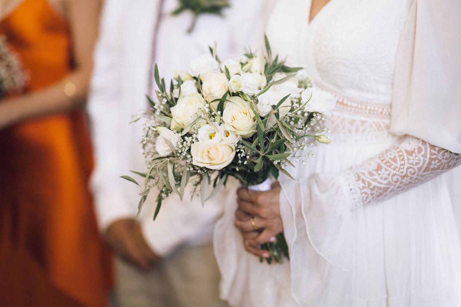 Bride in white lace gown holding elegant bouquet of white roses and greenery at outdoor wedding ceremony.