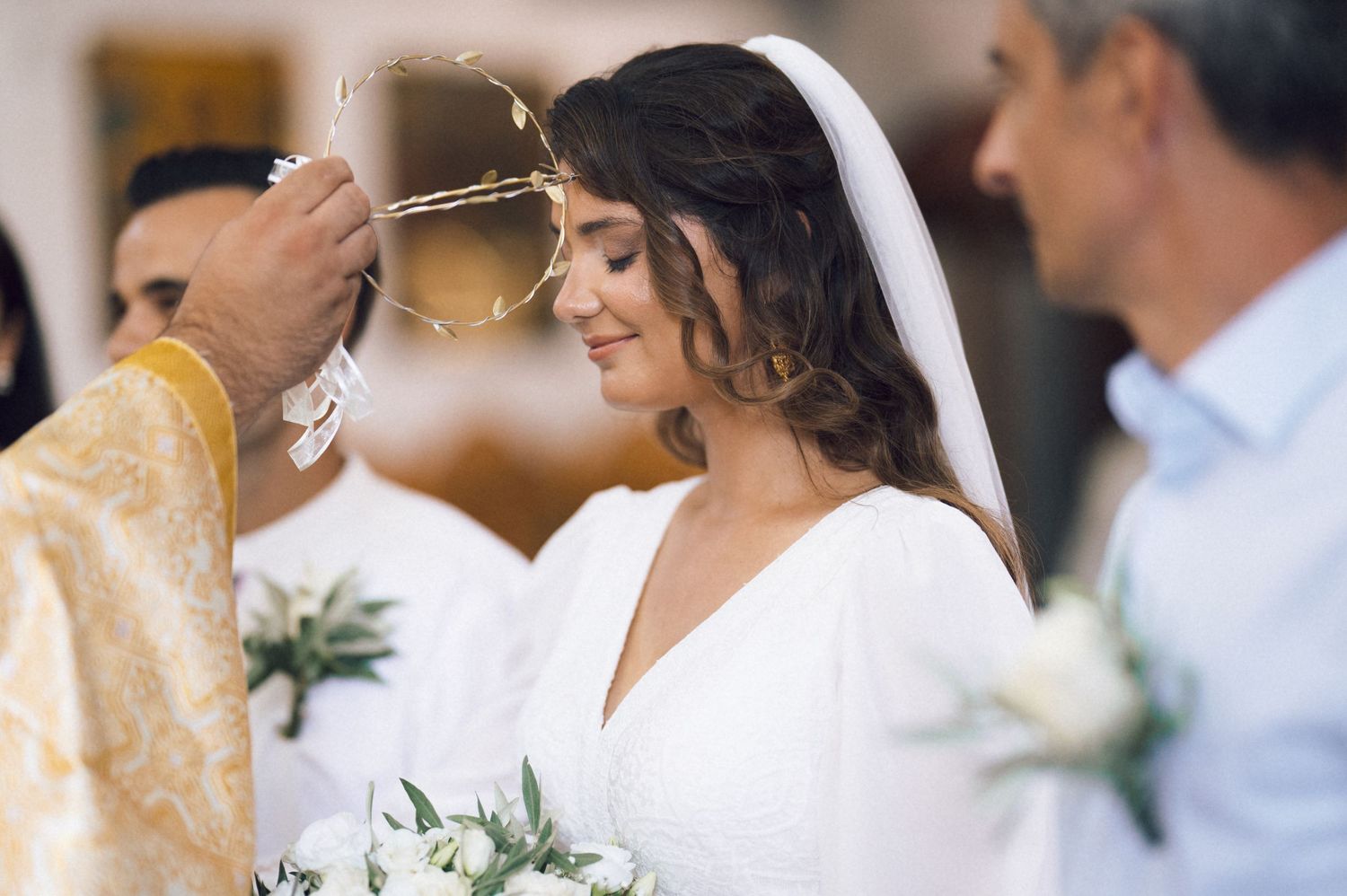 Bride in white dress and veil receiving blessing during Orthodox wedding ceremony, holding bouquet.