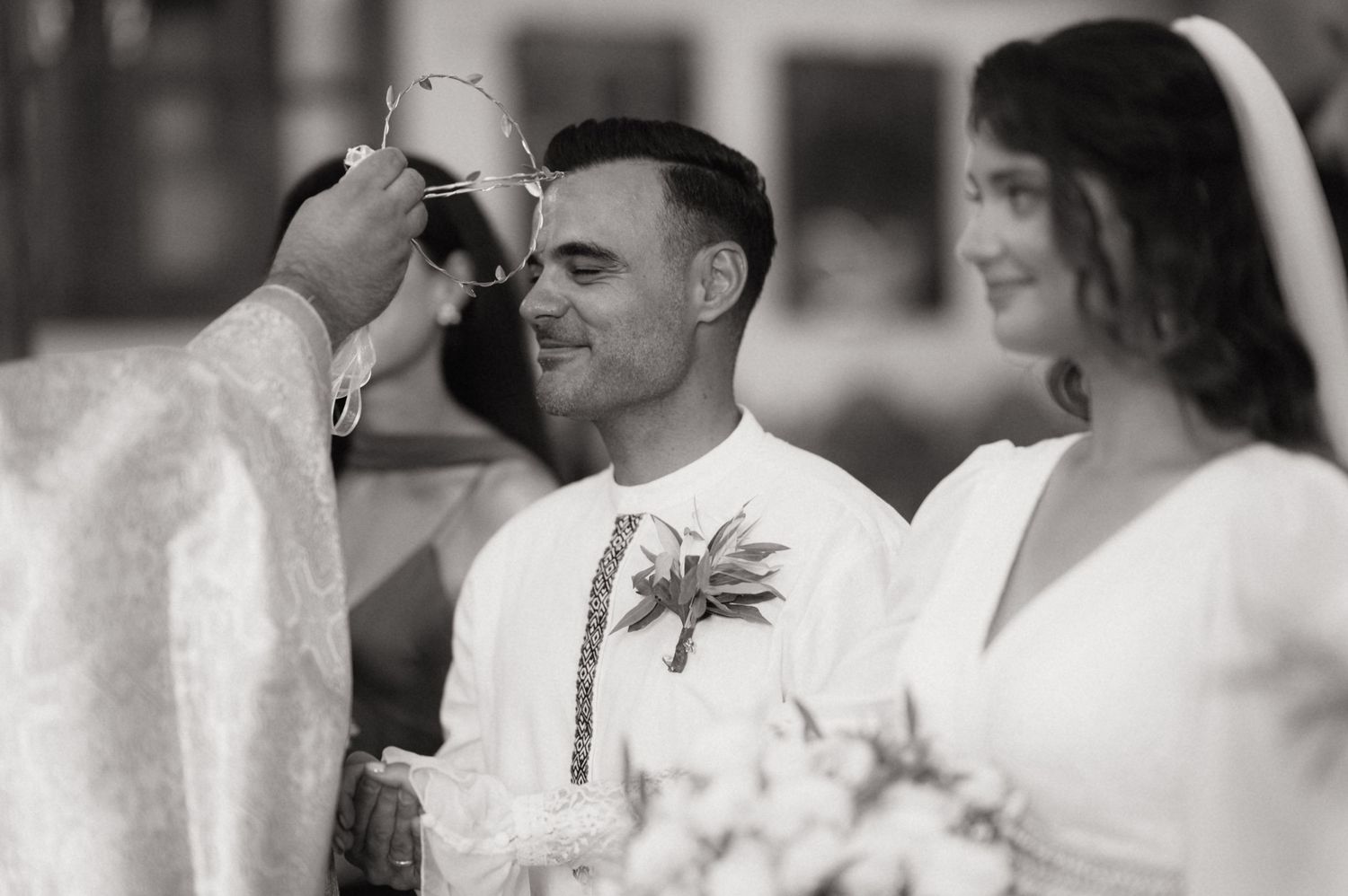 Black and white wedding ceremony photo of groom smiling as officiant performs blessing with bride holding bouquet.