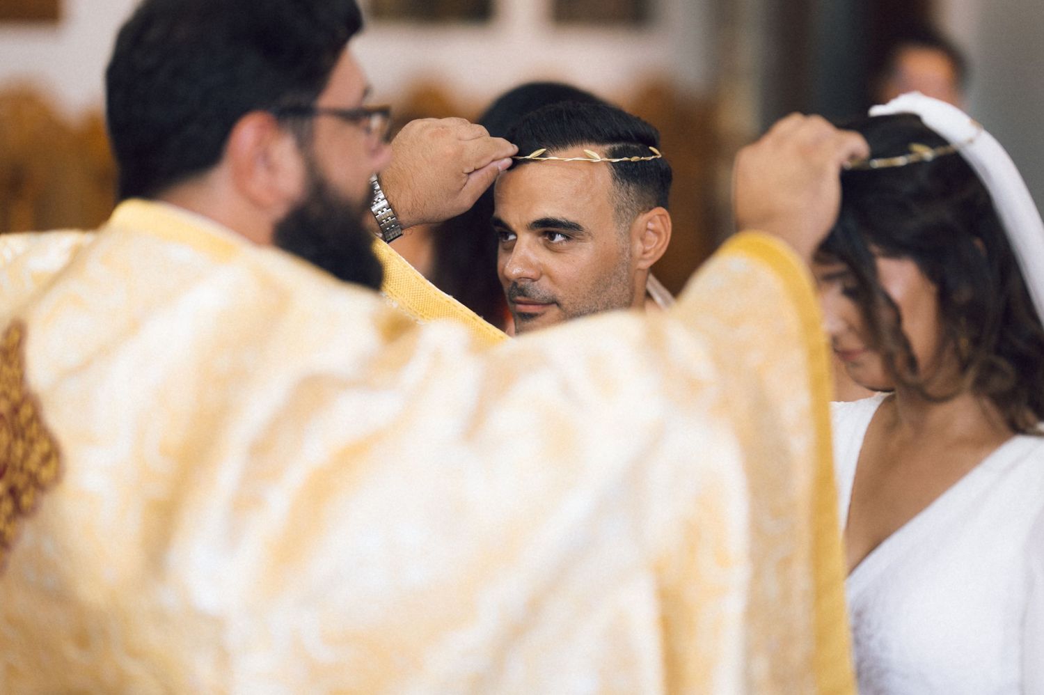 Orthodox priest placing crowns on couple during traditional religious wedding ceremony in golden vestments.