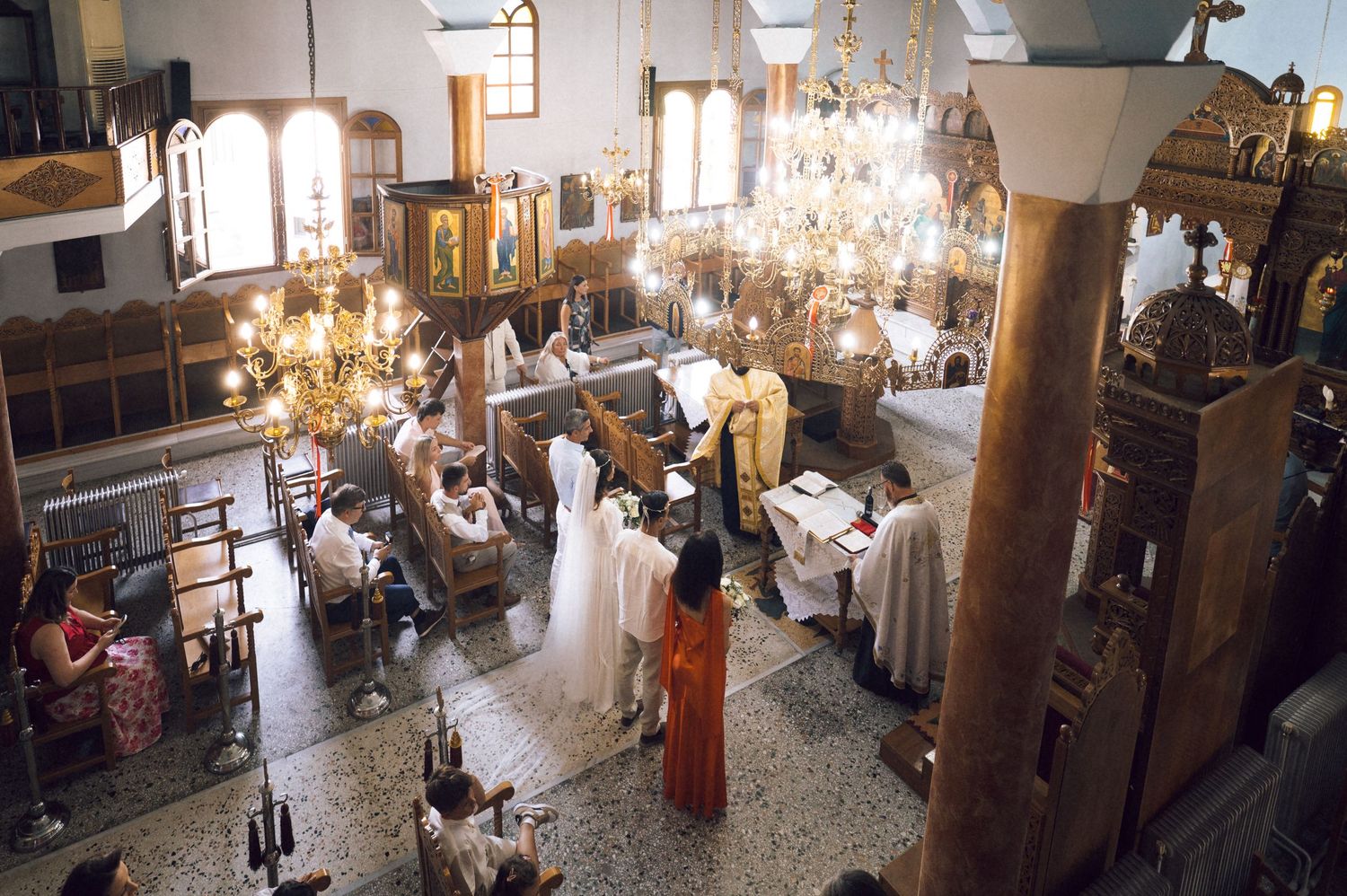 Orthodox church ceremony with clergy and congregation gathered under ornate chandeliers in a decorated sanctuary.