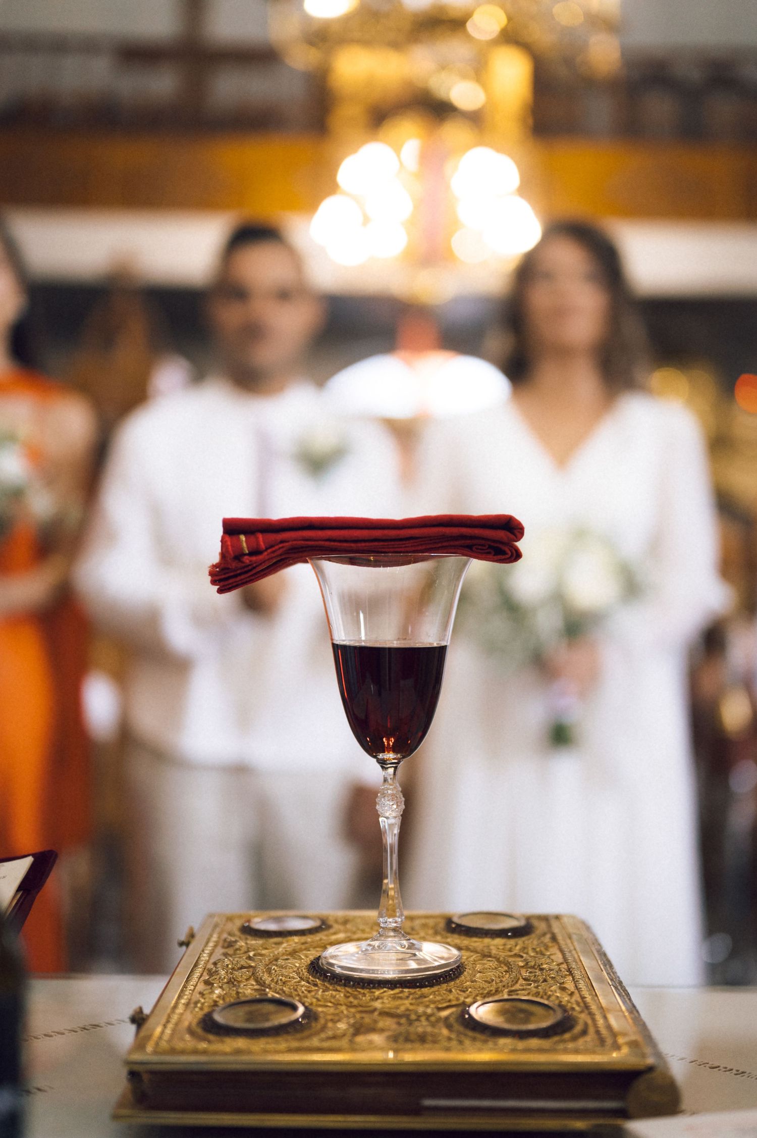 Orthodox wedding ceremony wine glass with red cloth on golden Bible, blurred bride and groom in white in background.