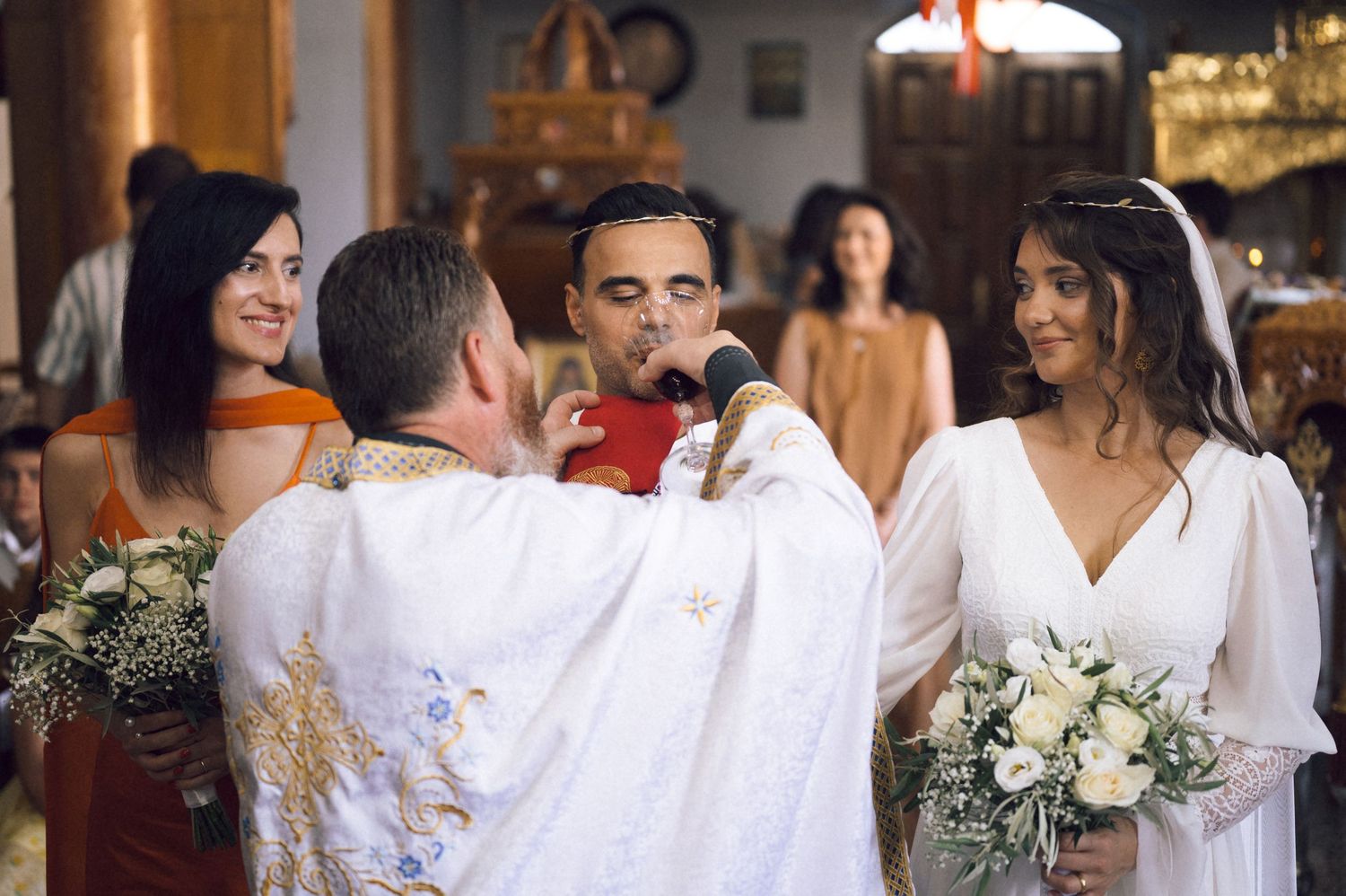 Orthodox wedding ceremony with bride in white gown, groom, and priest holding a chalice in a decorated church.