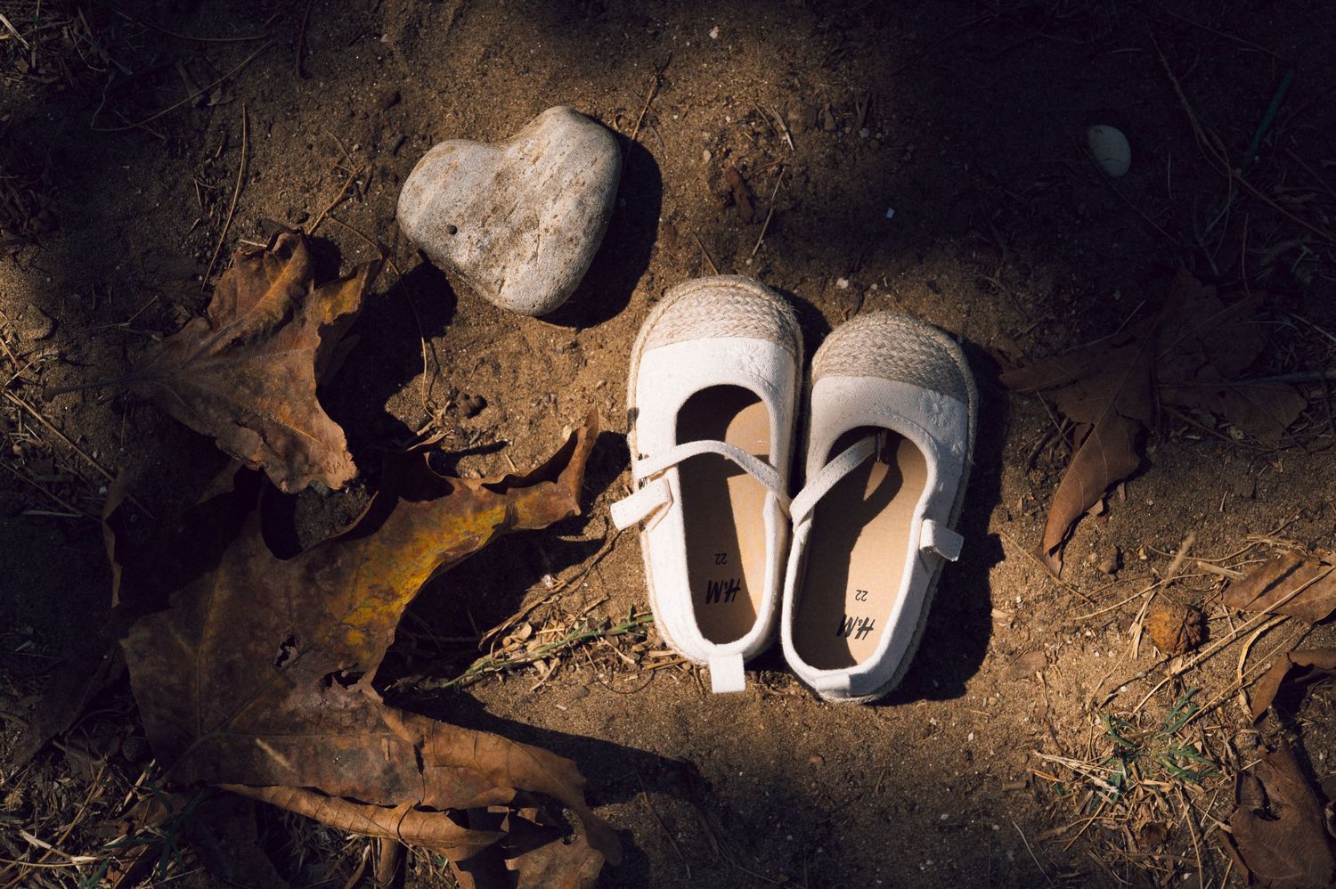 Small white children's shoes resting on dirt ground beside a rock and dry leaves in dim lighting.