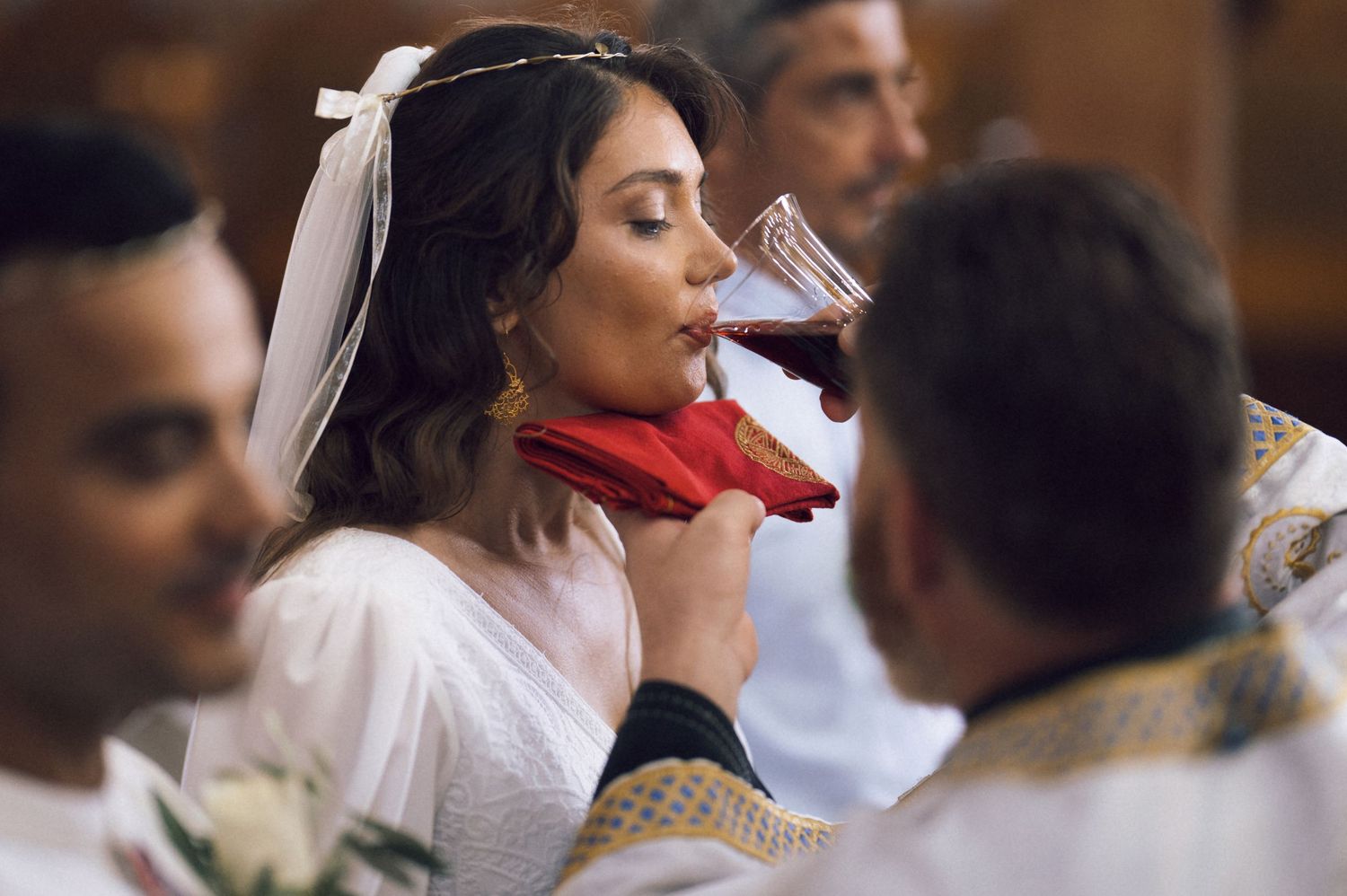 Bride in white dress and veil drinking red wine from a chalice during a religious wedding ceremony.