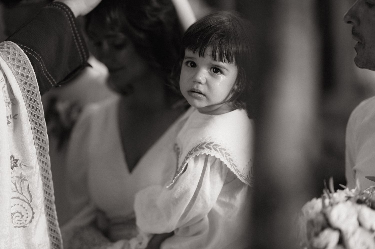 Black and white photo of a young girl in white dress being held at a wedding ceremony.