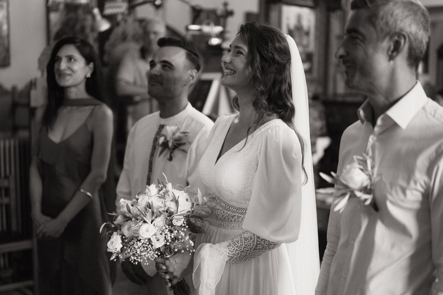 Black and white wedding photo of bride holding bouquet with groom and guests smiling during ceremony.