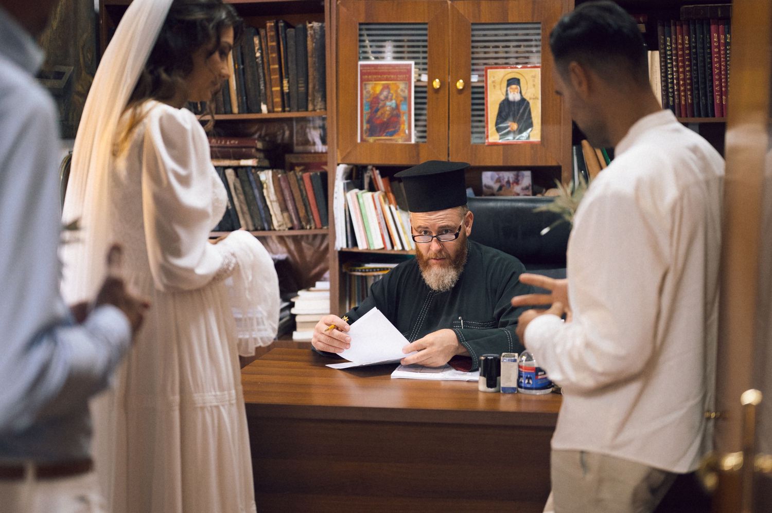 Orthodox priest in black vestments reviews documents at desk surrounded by books and visitors in a church office.