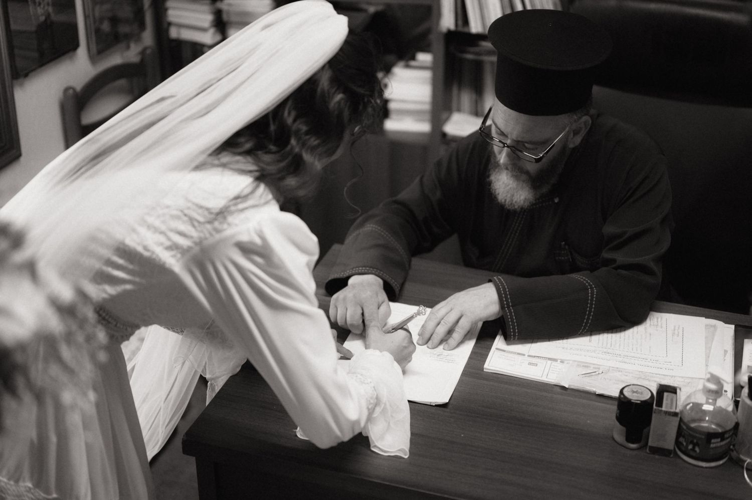 Black and white photo of a bride in white veil signing documents with an orthodox priest at a desk.