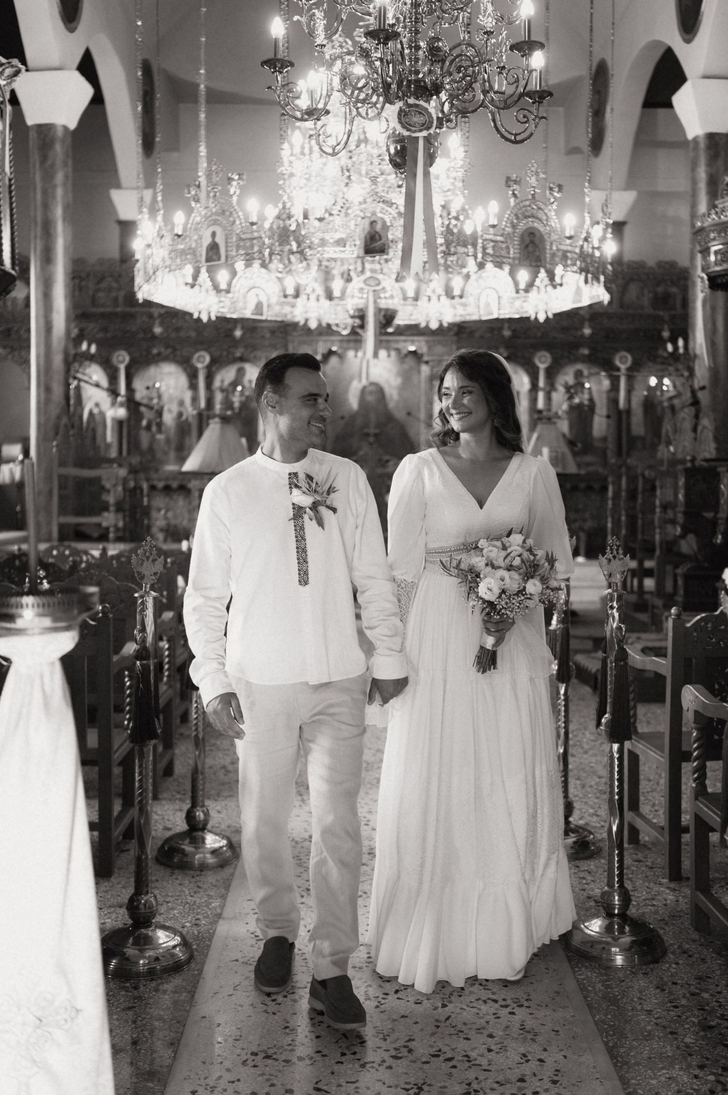Black and white photo of a couple in wedding attire walking hand in hand down a church aisle under a chandelier.