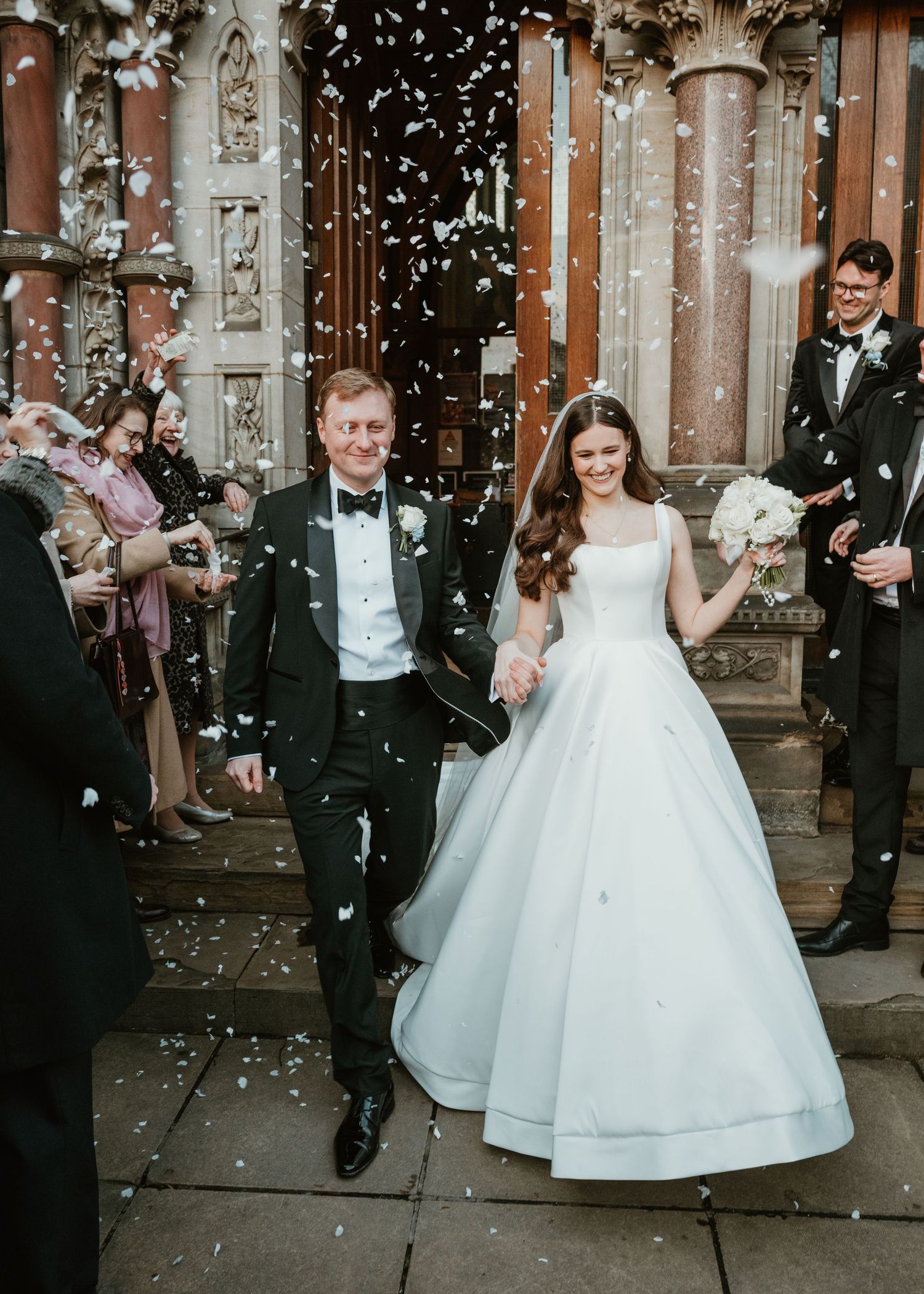 Bride and groom exit church amid confetti shower, she in white ballgown with bouquet, he in black tuxedo.