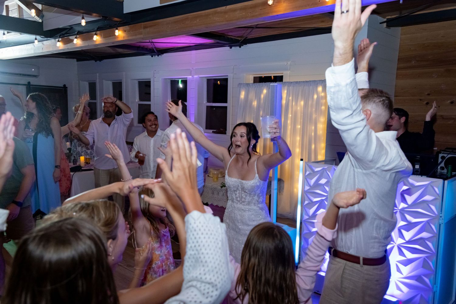 Bride and guests dancing joyfully at a wedding reception with colorful lights and hands raised.