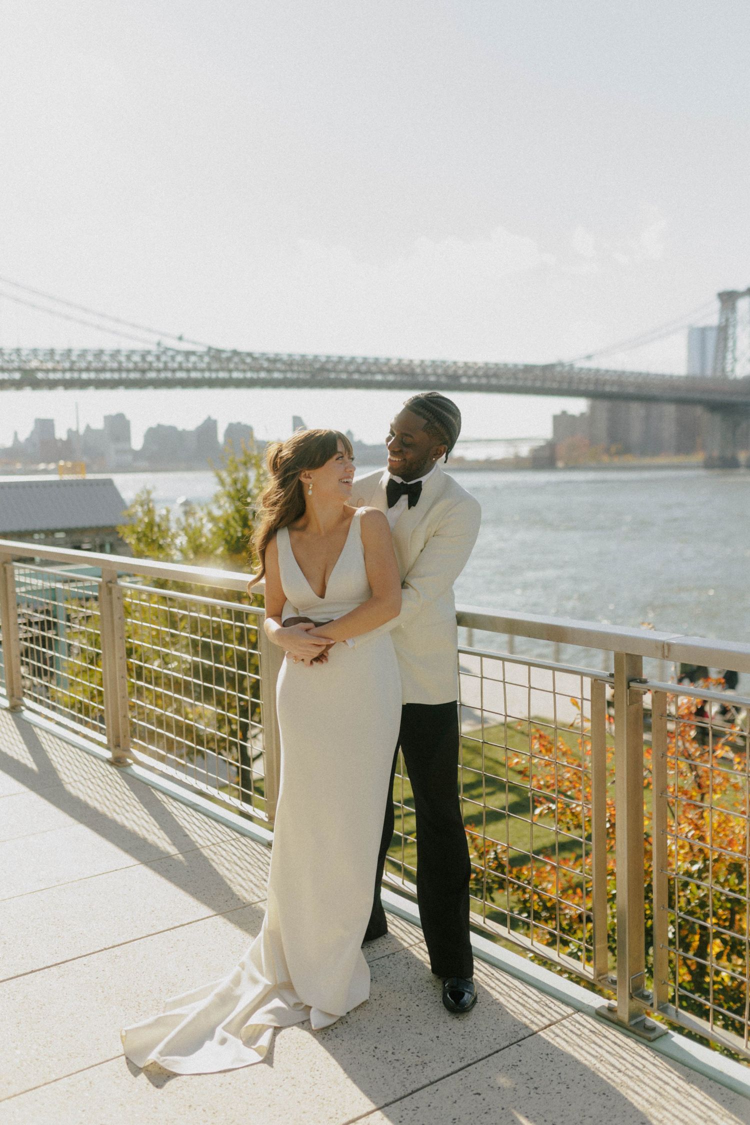 Couple in wedding attire embracing on a waterfront terrace with the Brooklyn Bridge in the background.