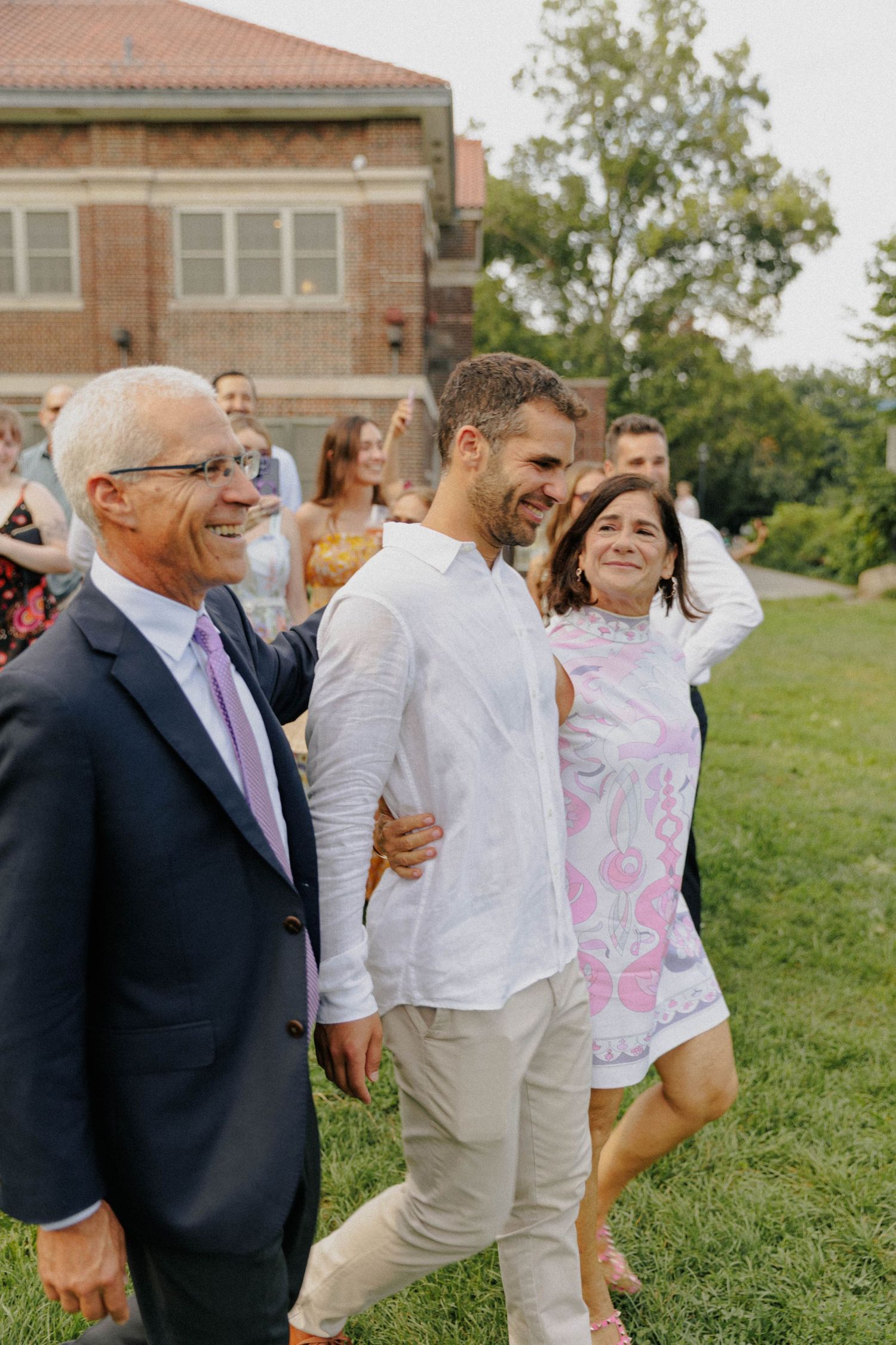 A man in a white shirt walks with smiling family members outdoors near a brick building at a wedding celebration.