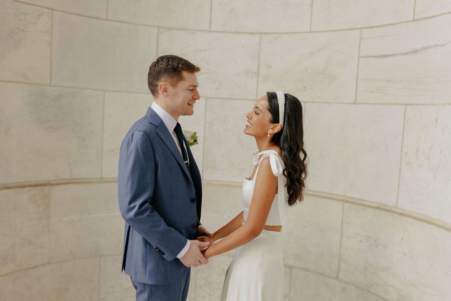 A bride and groom hold hands and smile at each other in a white marble venue on their wedding day.