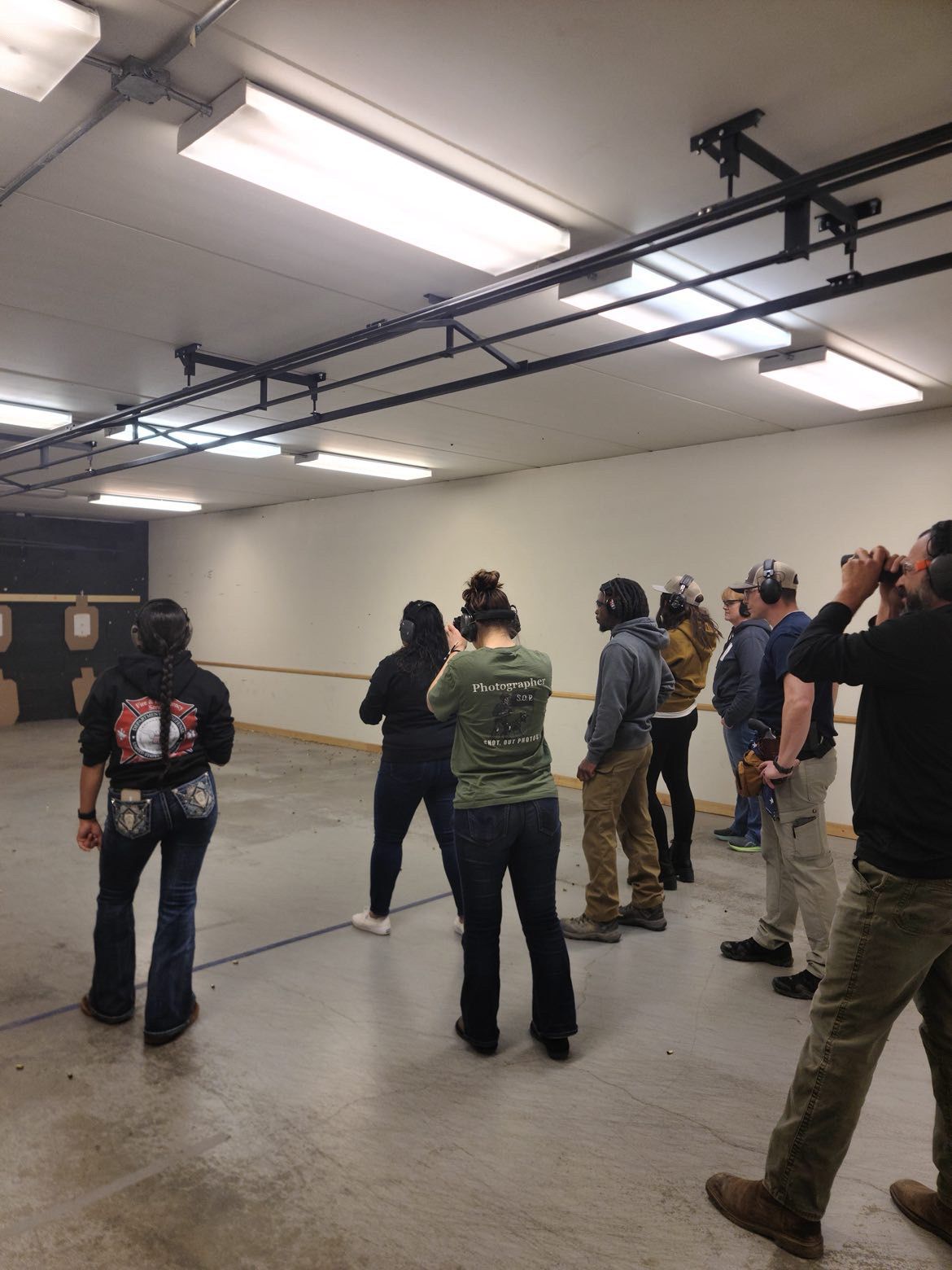 Group of people standing at an indoor shooting range with targets on the wall under fluorescent lights.